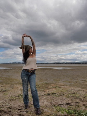 A woman stands on a muddy shoreline under a dramatic cloudy sky, stretching her arms overhead. She is wearing a beige knitted corset top, blue jeans, and dark boots.
